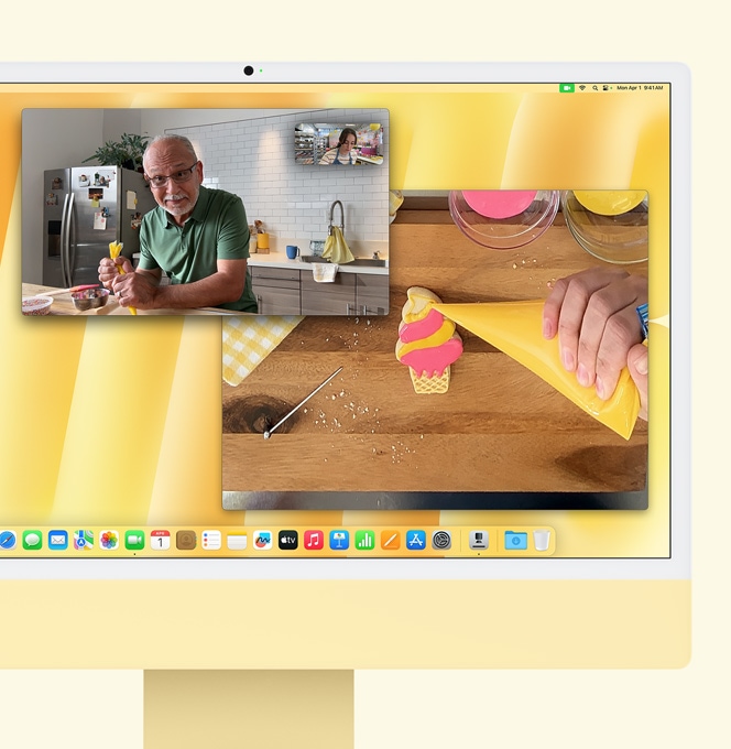 Geltonos spalvos „iMac“. „Desk View“ fukcijos demonstracija. Ekrane rodomas kepėjas, kuris kalbasi vaizdo skambučiu su kitu asmeniu. Viename ekrane rodomas kepėjo ir vyro veidas. Kitame ekrane rodomos kepėjo rankos, glazūruojančios sausainį.