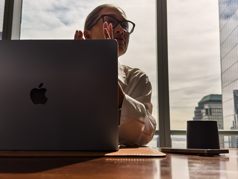 Yüksek katlı bir binadaki masada oturan bir kadın. Arkasında diğer binaları gösteren büyük bir pencere var. Kadının önünde bir MacBook açık.