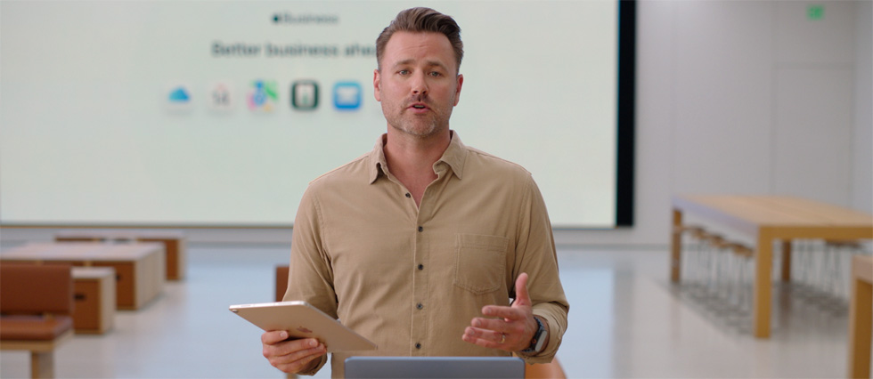 El presentador habla mientras hace referencia a un iPad, y al fondo se muestra una presentación y varios íconos de apps para empresas en una pantalla grande.