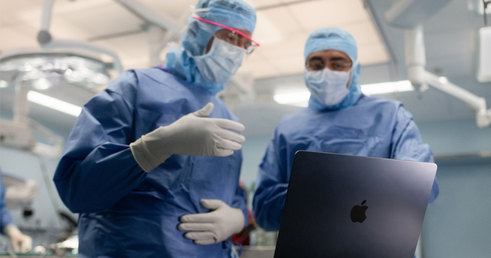 Hai bác sĩ phẫu thuật trong phòng mổ nhìn vào MacBook, một người ra hiệu bằng tay trong khi người kia quan sát