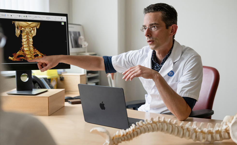 Un profesional médico señala una representación en 3D de la parte trasera del cuello en un Apple Studio Display, con un MacBook sobre la mesa