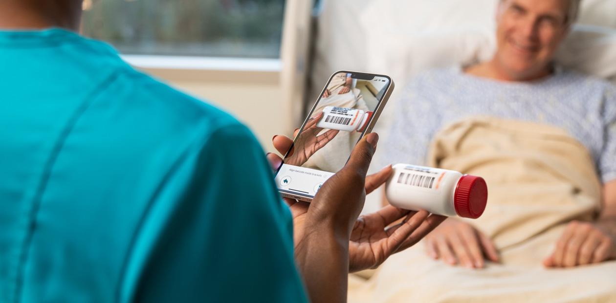 Enfermeira com uniforme azulado escaneia o medicamento de um paciente usando um iPhone.