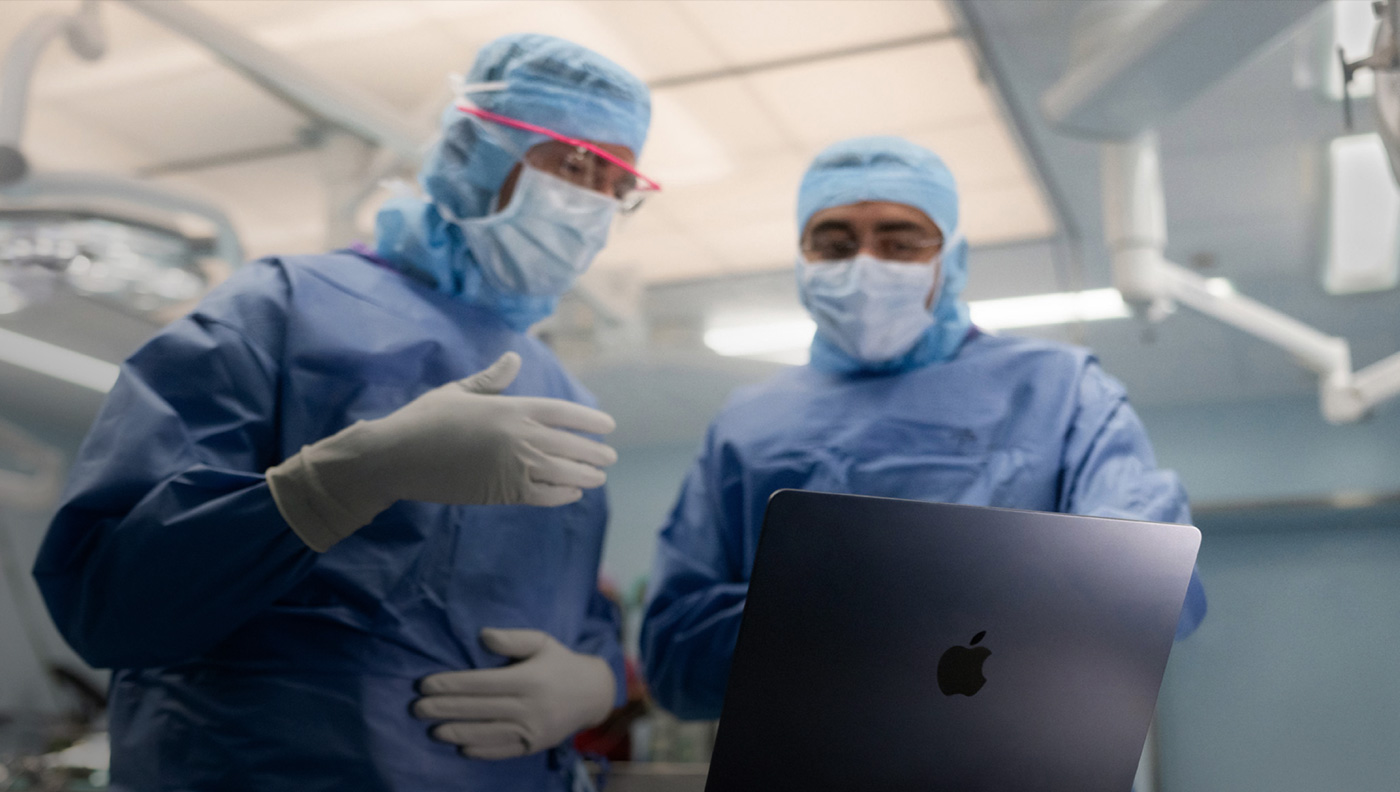 Dois cirurgiões conversando sobre algo na tela de um MacBook Pro na sala de cirurgia.