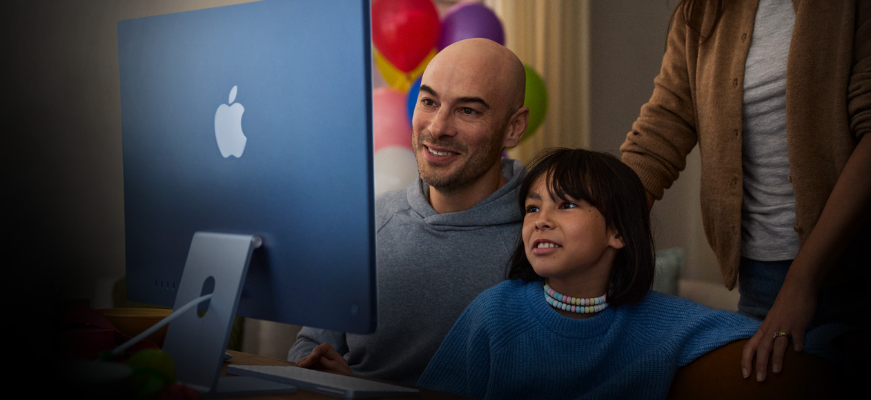 شخصان يجلسان معاً ويستخدمان جهاز iMac بينما يقف شخص ثالث خلفهما ويراقبهما.