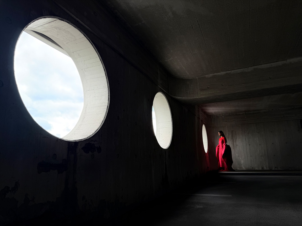 Eine Frau in einem langen roten Kleid steht in einem leeren Raum und wird fotografiert. Die Fotos App wechselt durch die Brennweiten, um die Möglichkeiten des Pro Kamera-Systems zu zeigen, hier: 24 mm